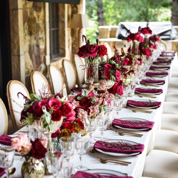 Elegant long dining table with red floral arrangements and neatly set with glassware and napkins.