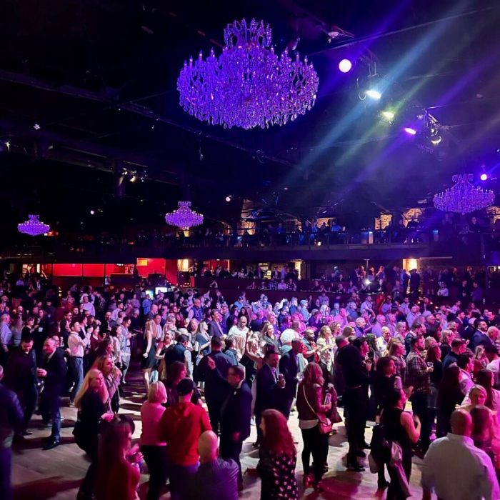 Crowded ballroom with dancing people under large chandeliers and colorful lights.